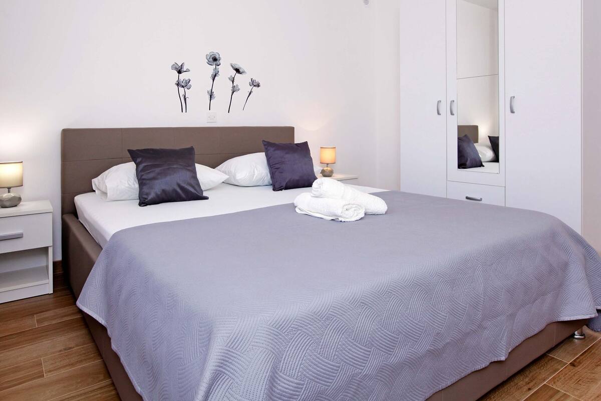 Una cama bien tendida con una colcha gris y dos almohadas, una mesita de noche con una lámpara, una obra de arte en la pared y un armario con espejo.