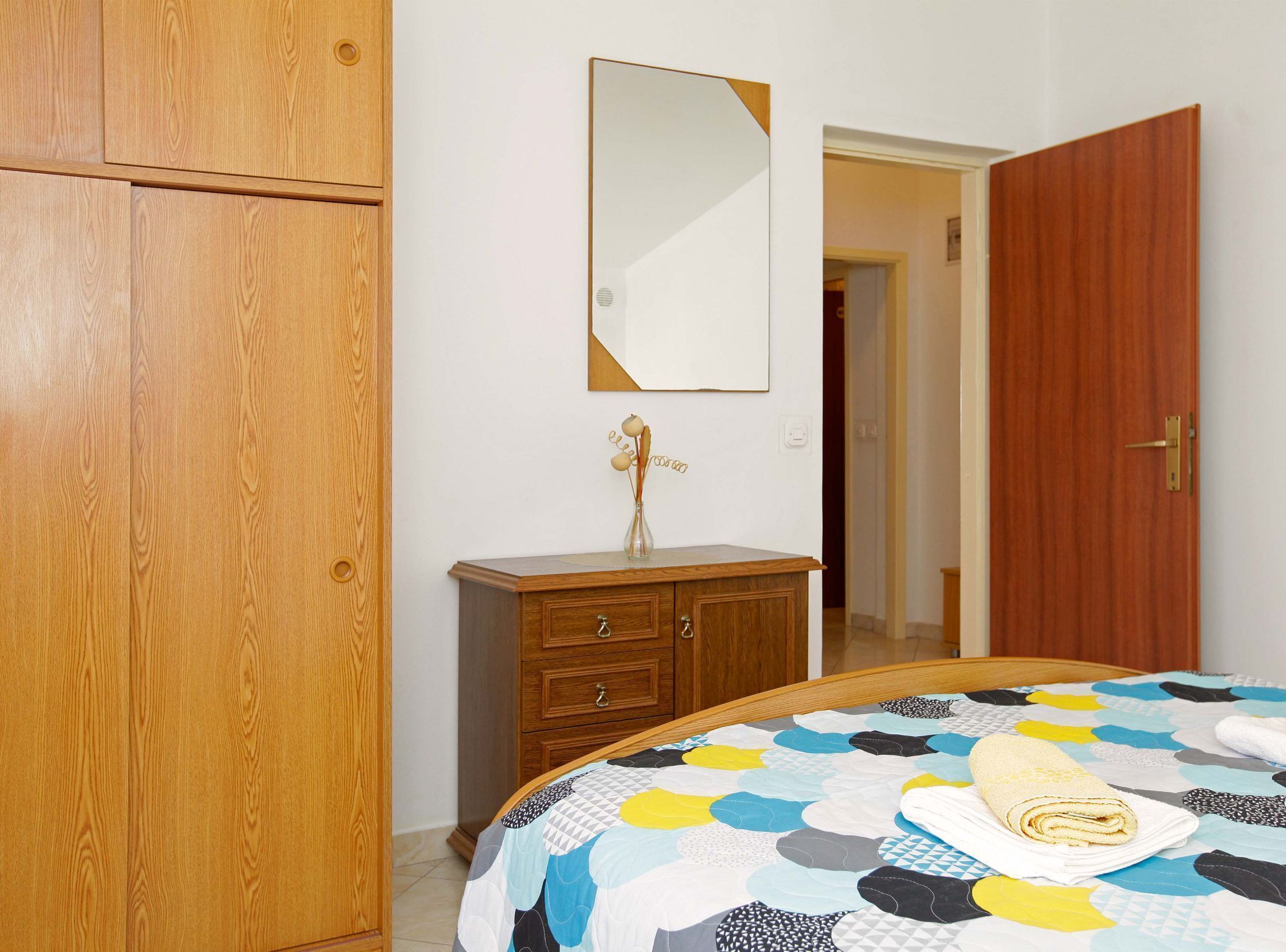 Un dormitorio con un armario de madera, un aparador, un espejo y una cama con colcha colorida.