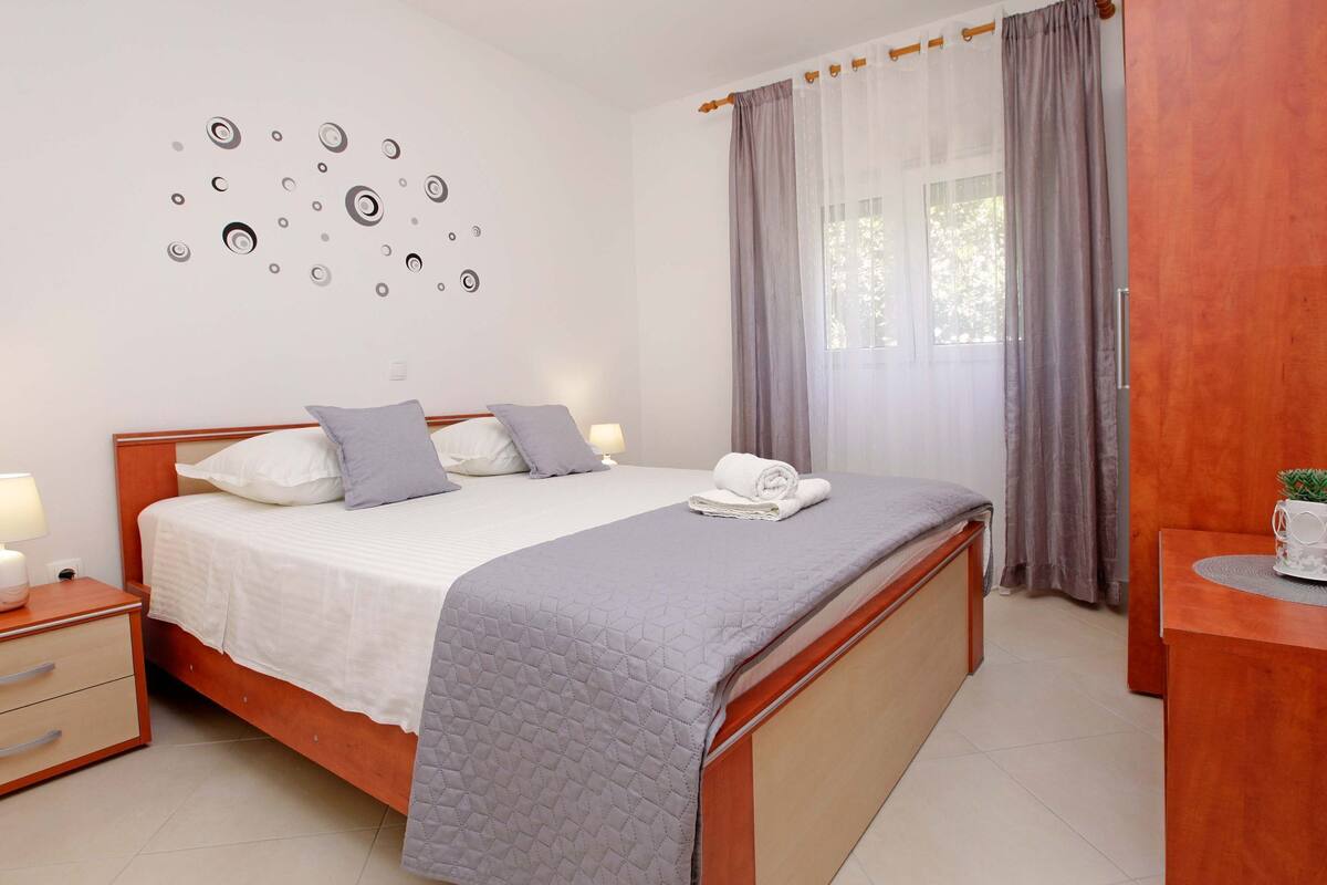 Una cama bien hecha con colcha gris, una mesita de noche con lámpara, un reloj de pared y una pequeña mesa con una planta.