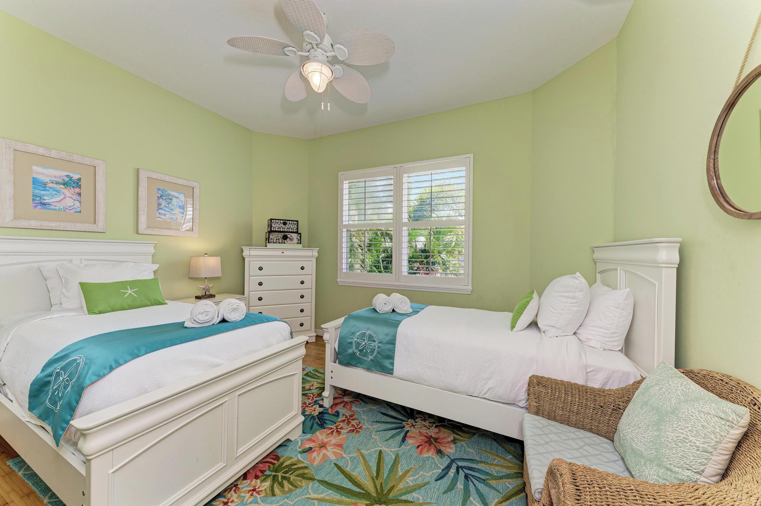 A bedroom with two beds, a ceiling fan, a window with blinds, and a wicker chair.