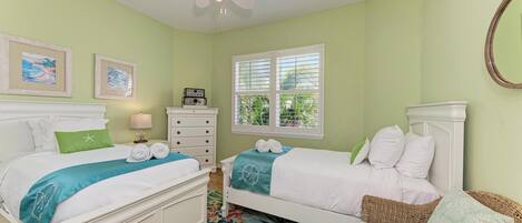 A bedroom with two beds, a ceiling fan, a window with blinds, and a wicker chair.