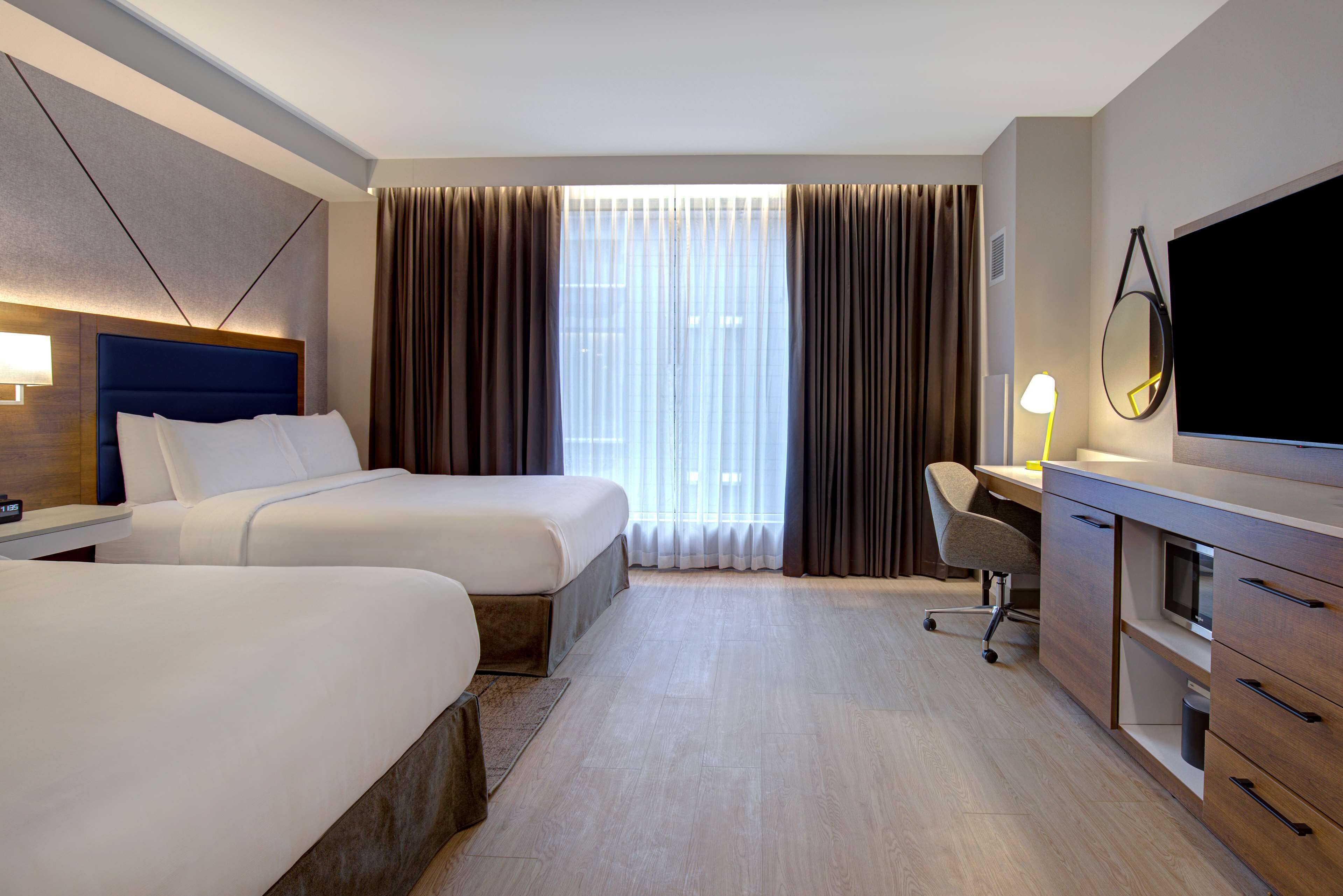 A hotel room with two beds, a desk, a chair, a TV, and a large window with curtains.