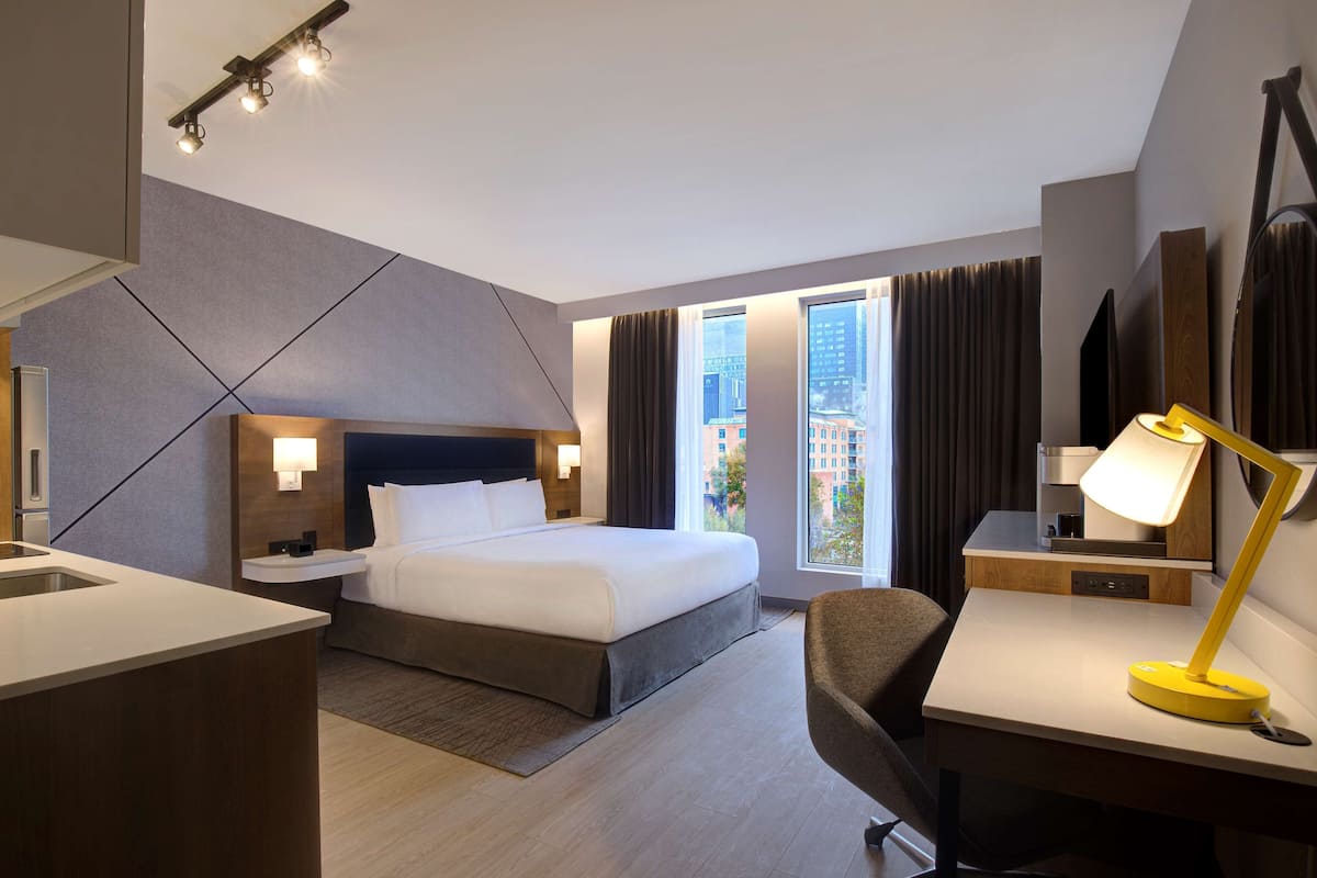 A modern hotel room with a large bed, a desk, a chair, and a view of the city through large windows.