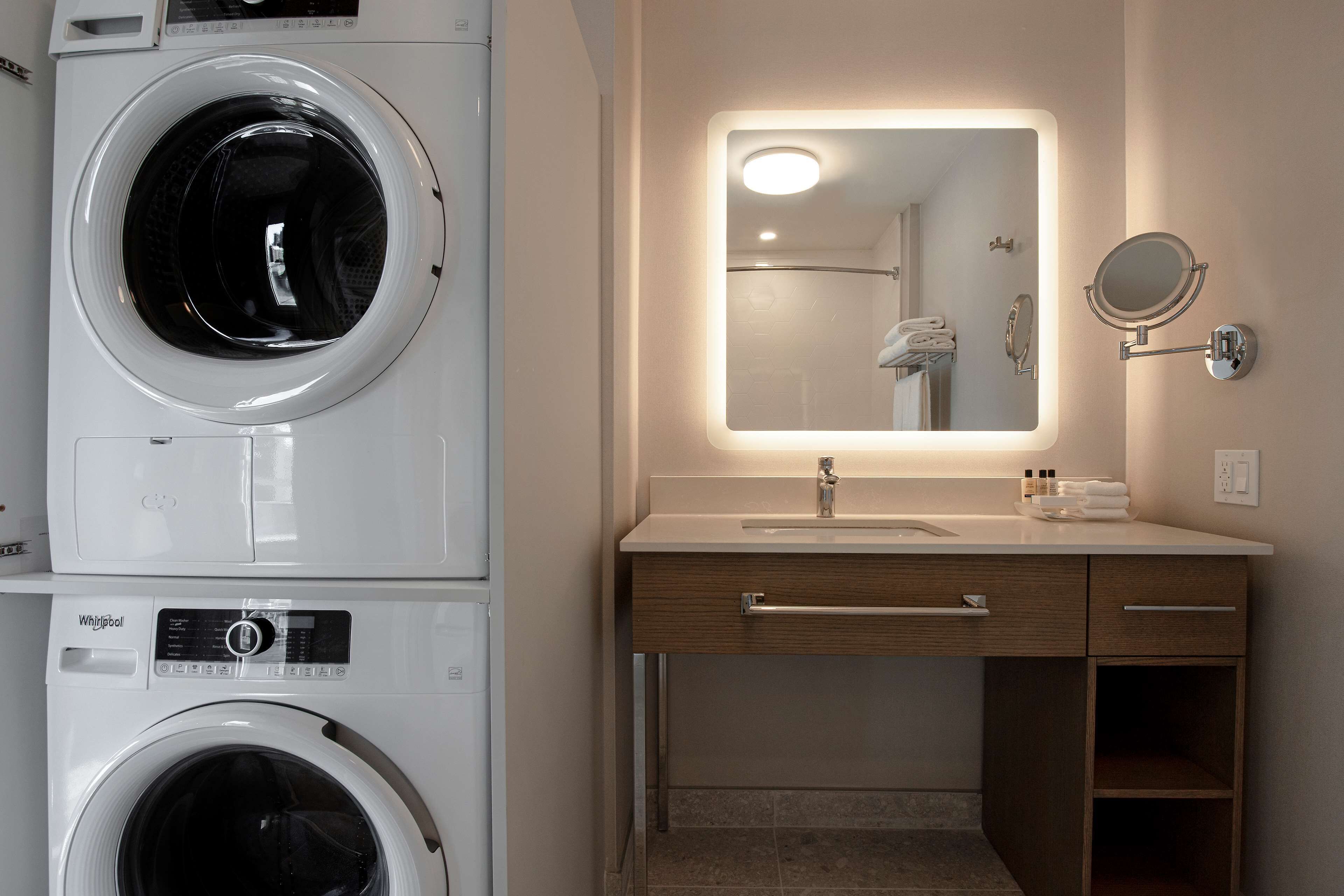 A hotel bathroom with a stacked washer and dryer, a vanity with a mirror, and a shower area.