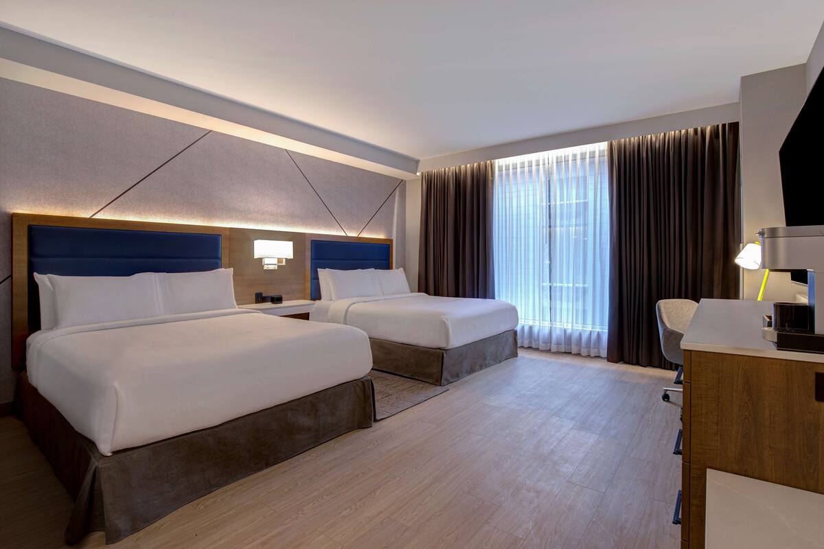 A hotel room with two beds, a desk, and a large window with curtains.