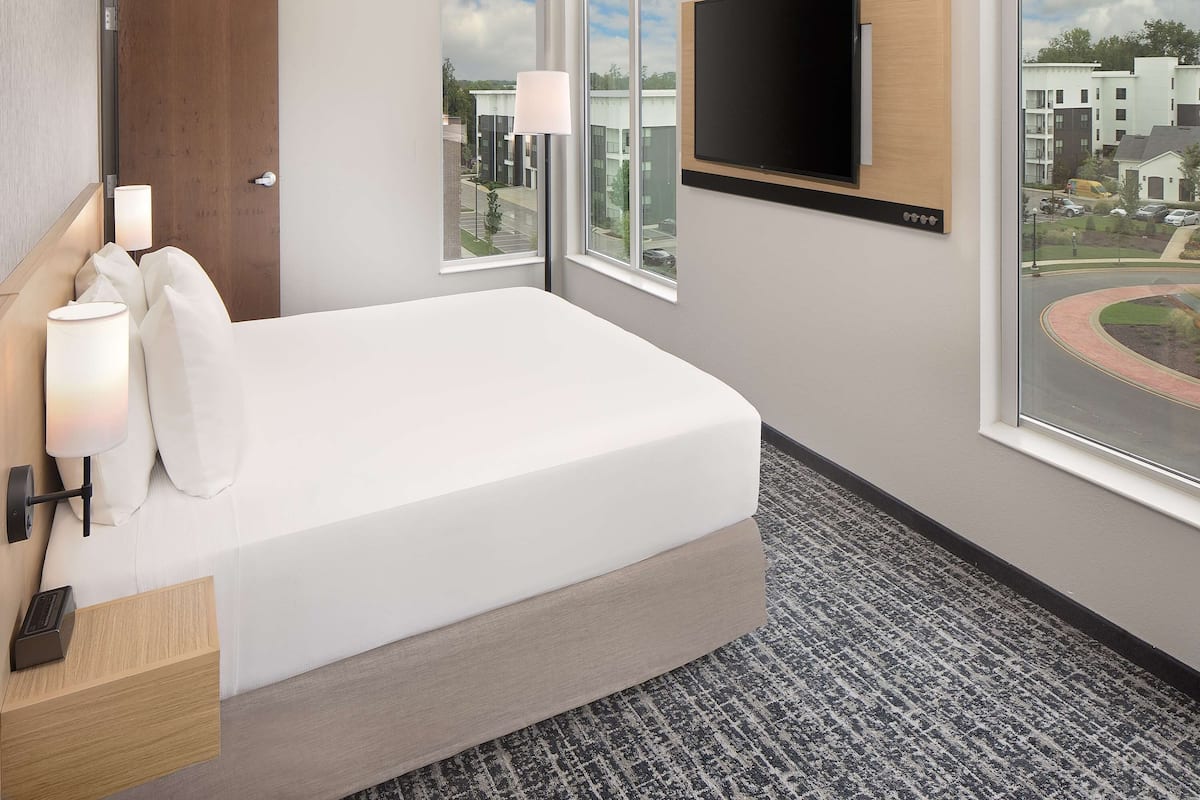 Quarto de hotel com uma cama, televisão na parede, secretária com luminária e janelas amplas com vista para a cidade.