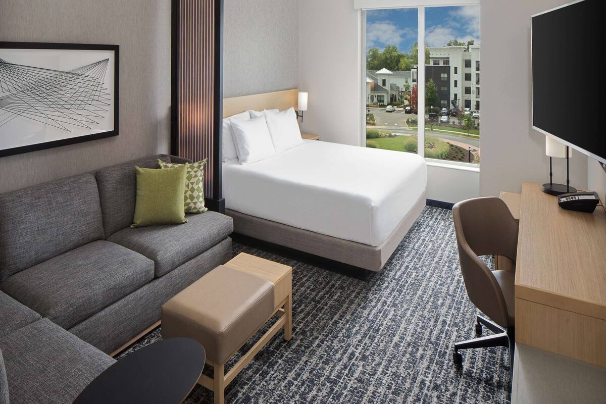 Quarto de hotel com cama, sofá, escrivaninha, cadeira e uma janela grande que oferece vista para prédios e árvores.