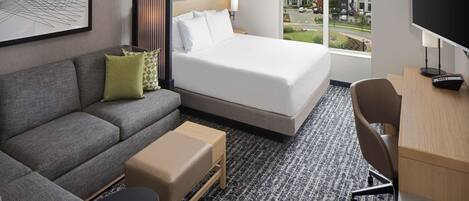 Une chambre d’hôtel comprenant un lit, un canapé, un bureau, une chaise et une grande fenêtre donnant sur des immeubles et des arbres.