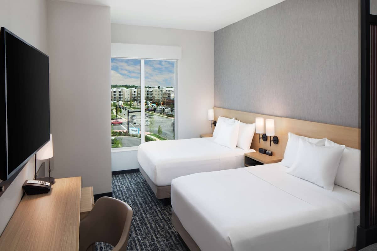 Quarto de hotel com duas camas, uma mesa de trabalho com uma televisão e vista da cidade por grandes janelas.