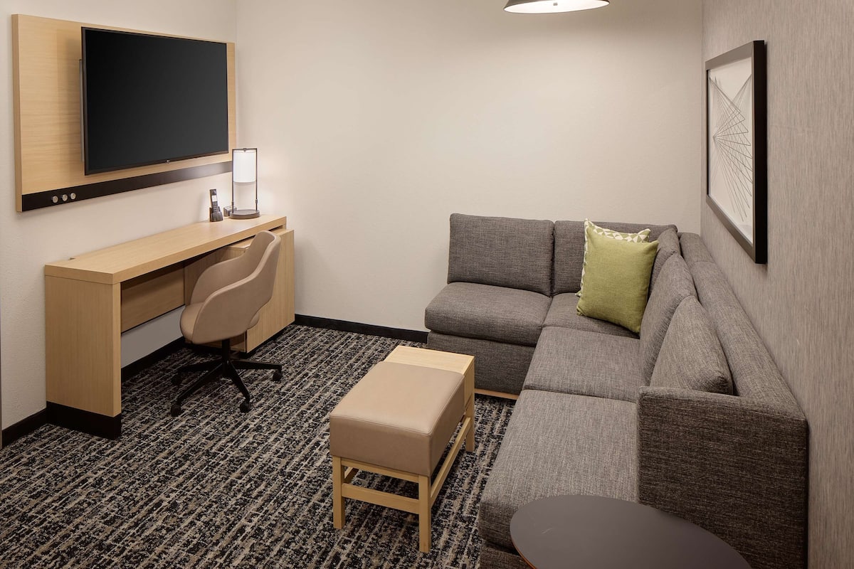 Quarto de hotel moderno com televisão de tela plana, escrivaninha com cadeira, sofá cinza com almofada verde, mesa redonda pequena e luminária pendente preta.