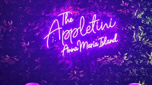 Een neonbord met de tekst 'The Appleletini Anna Maria Island', met twee witte stoelen en een kleine tafel voor een achtergrond van begroeiing.