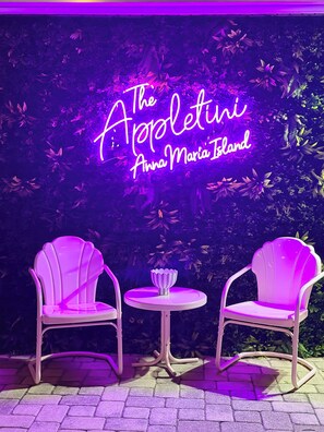 Neonvalomainos, jossa lukee ”The Appleletini Anna Maria Island”, ja sen edessä kaksi valkoista tuolia ja pieni pöytä, taustalla rehevää kasvillisuutta.