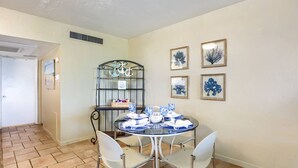 Un comedor con una mesa redonda dispuesta para cuatro personas, rodeada de sillas blancas. Hay un estante metálico con objetos decorativos y un cuadro en la pared.