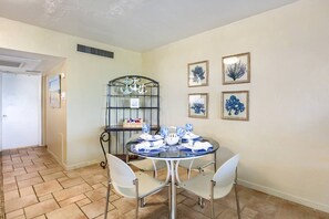 A dining area with a round table set for four, surrounded by white chairs. There is a metal shelf unit with decorative items and a framed picture on the wall.