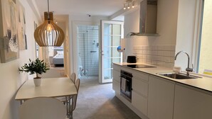 Une cuisine moderne avec un plan de travail blanc, un évier en acier inoxydable et un four encastré. On y trouve un coin repas avec une table et des chaises blanches, ainsi qu’une plante en pot posée sur la table.