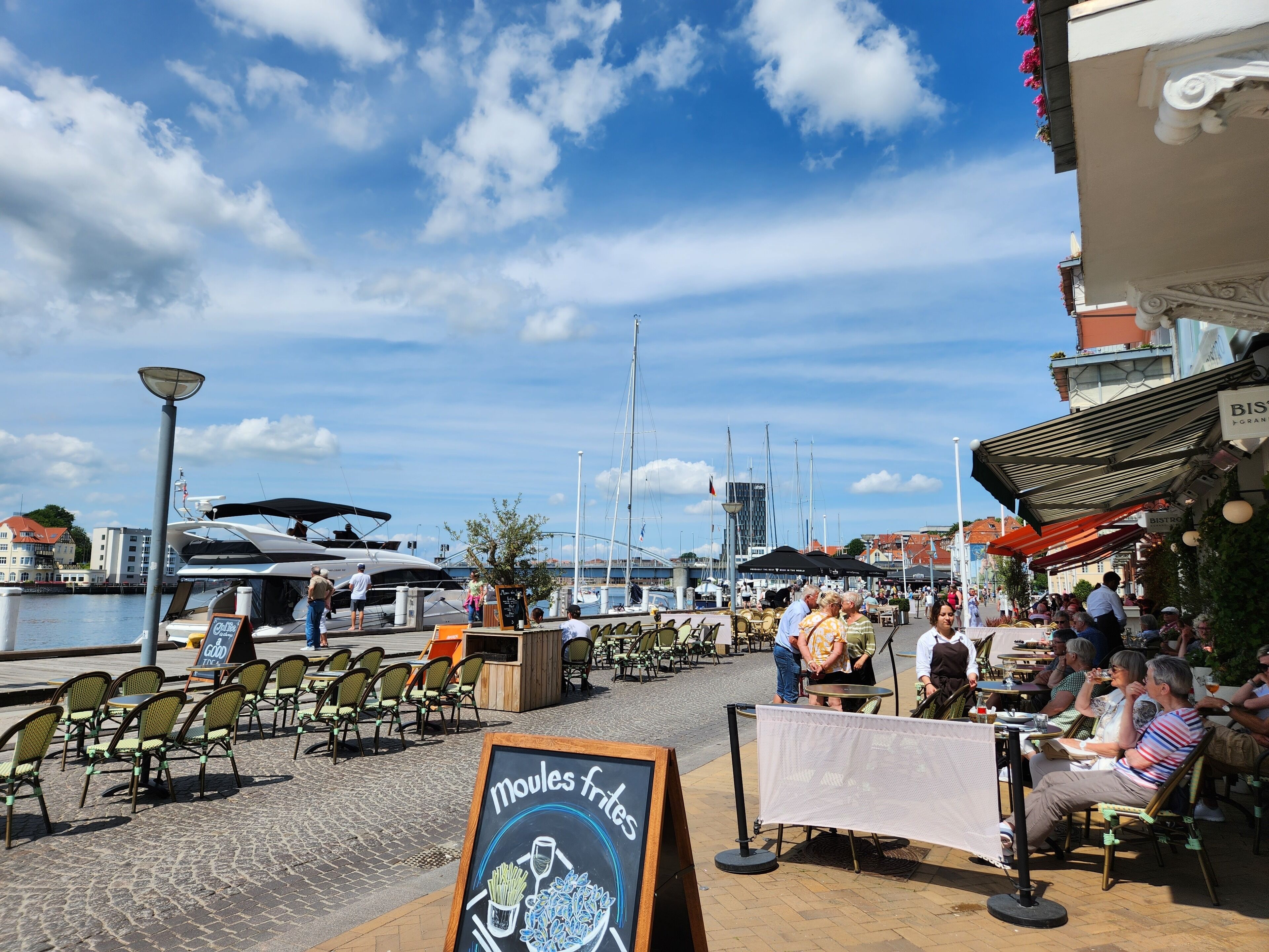 Ein Café am Wasser mit Außenbestuhlung, einem Schild für Moules-Frites und Blick auf Boote und Gebäude im Hintergrund.