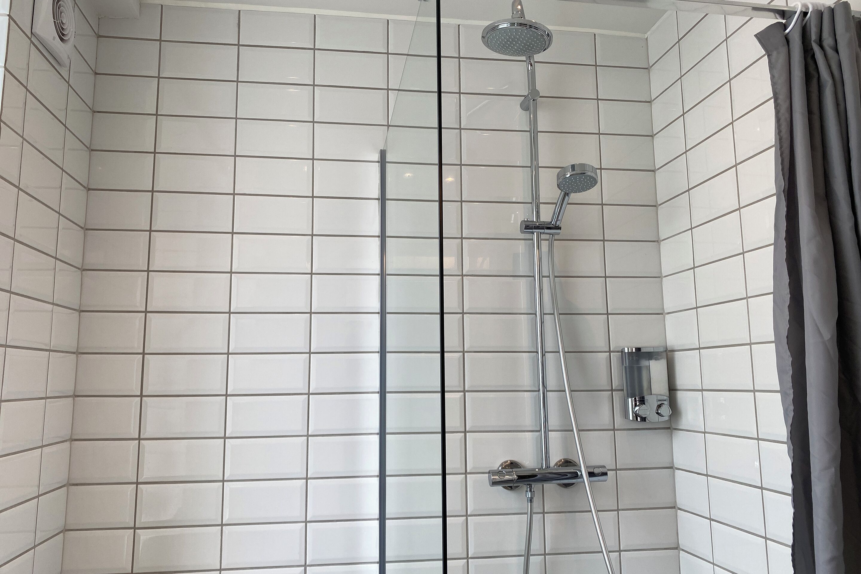 A shower with a handheld showerhead and a wall-mounted faucet, tiled walls, and a curtain.