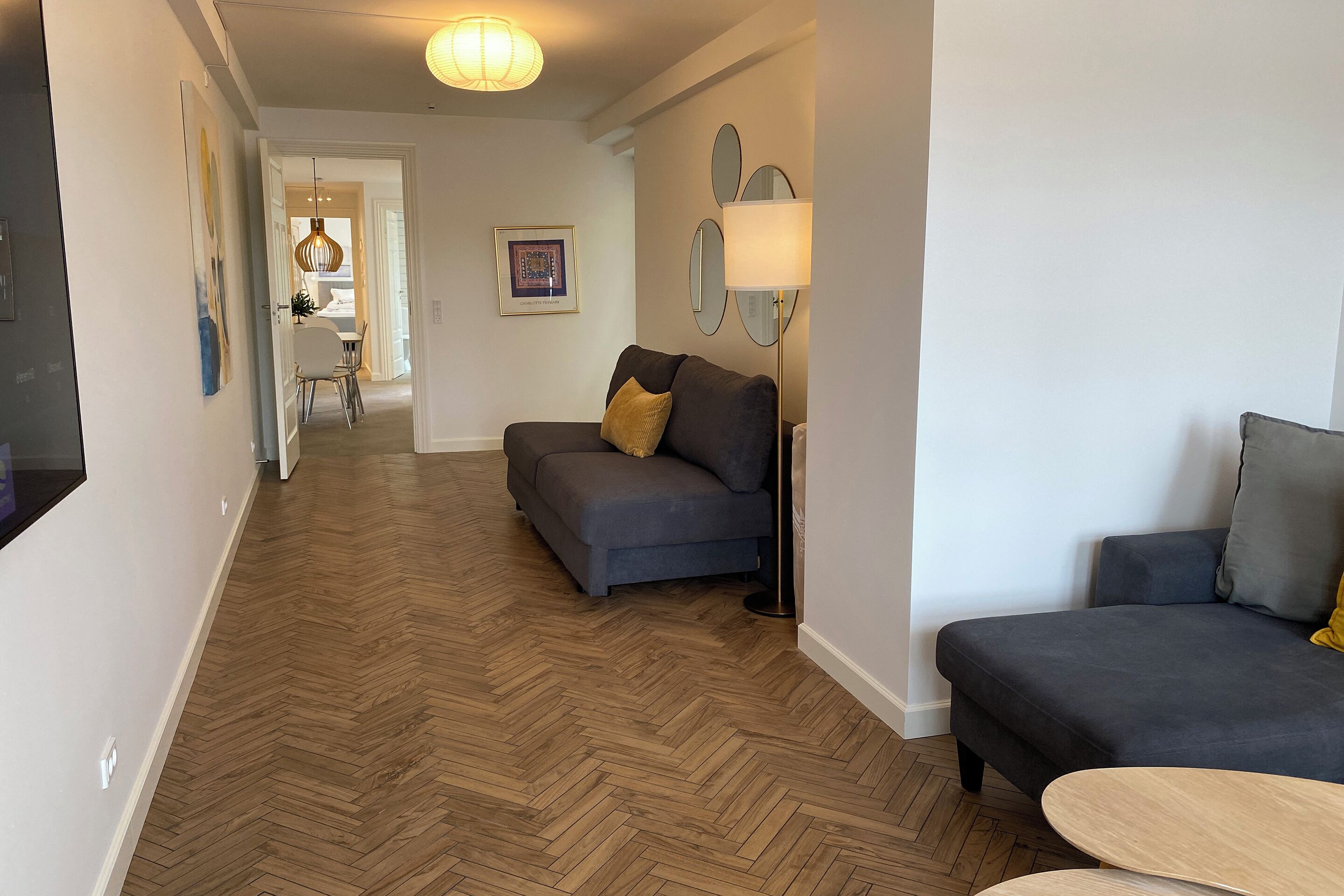 A modern living room with a herringbone wood floor, a dark grey sofa, a small round table, and a floor lamp.