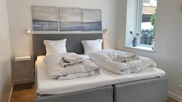 A neatly made bed with white linens and pillows, a grey headboard, and two wall-mounted artworks depicting a seascape.