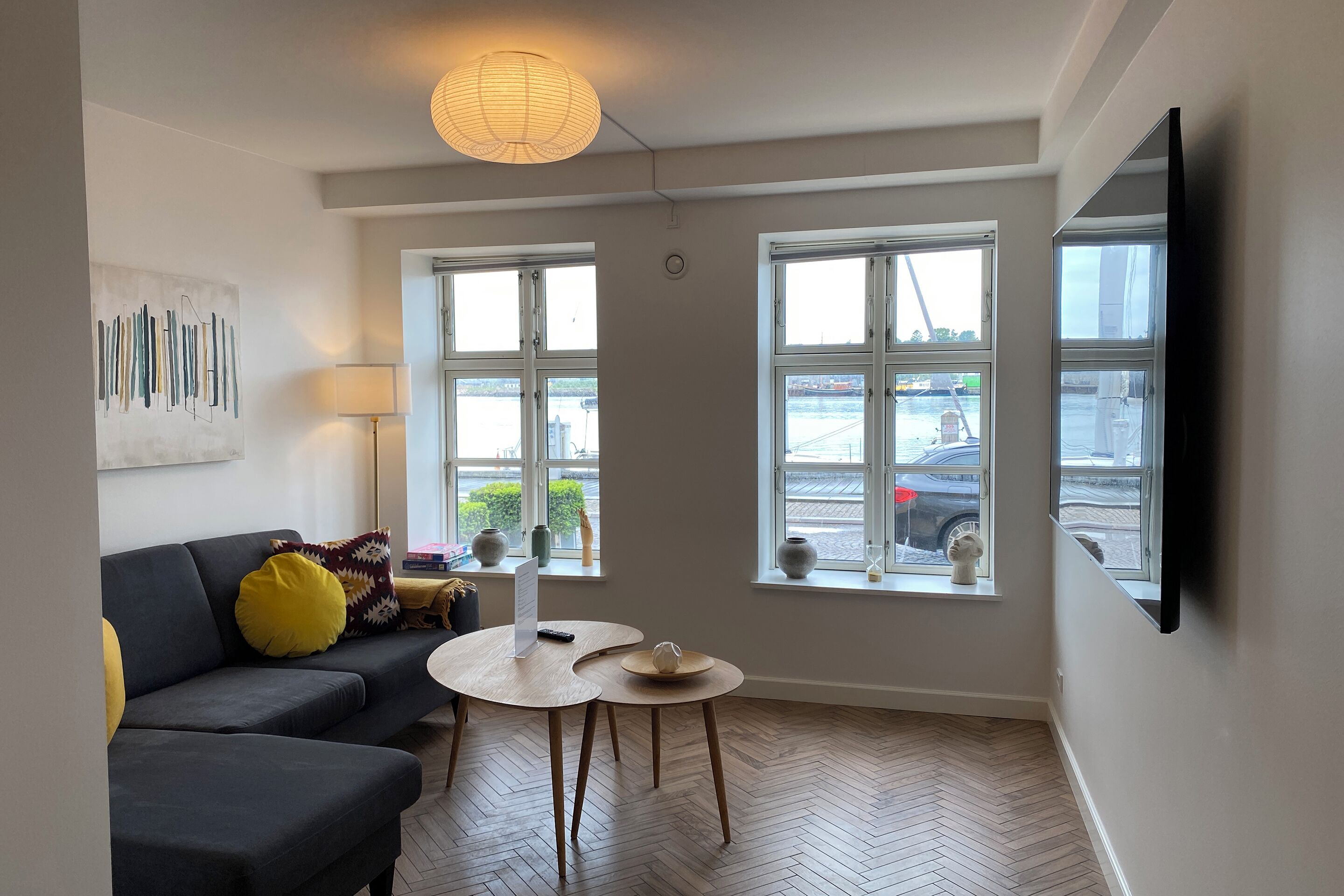 Ett modernt vardagsrum med en soffa, två små bord, en lampa och stora fönster med utsikt över staden.