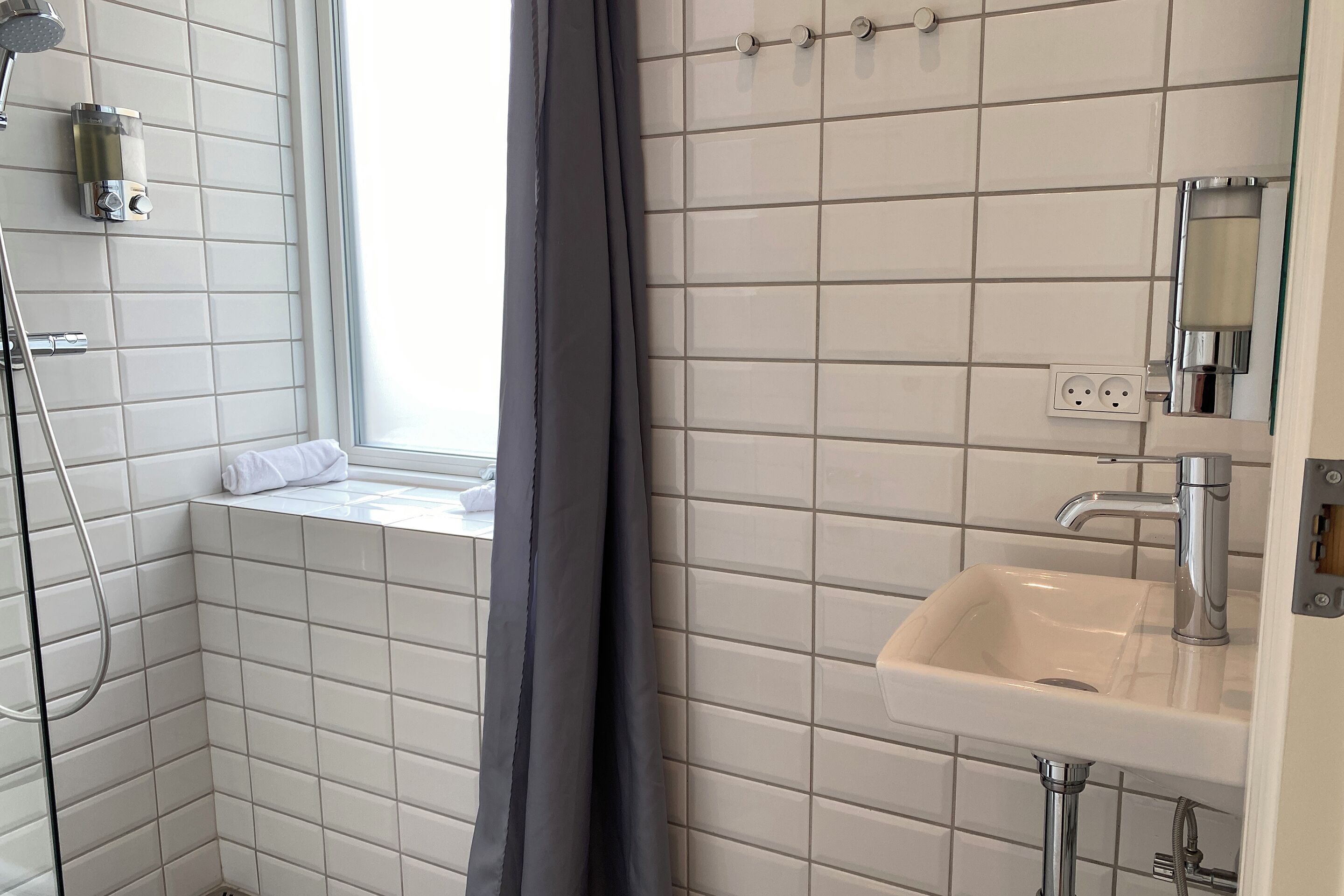 A bathroom with white tiles, a shower, a sink with a mirror, and a window with a towel.