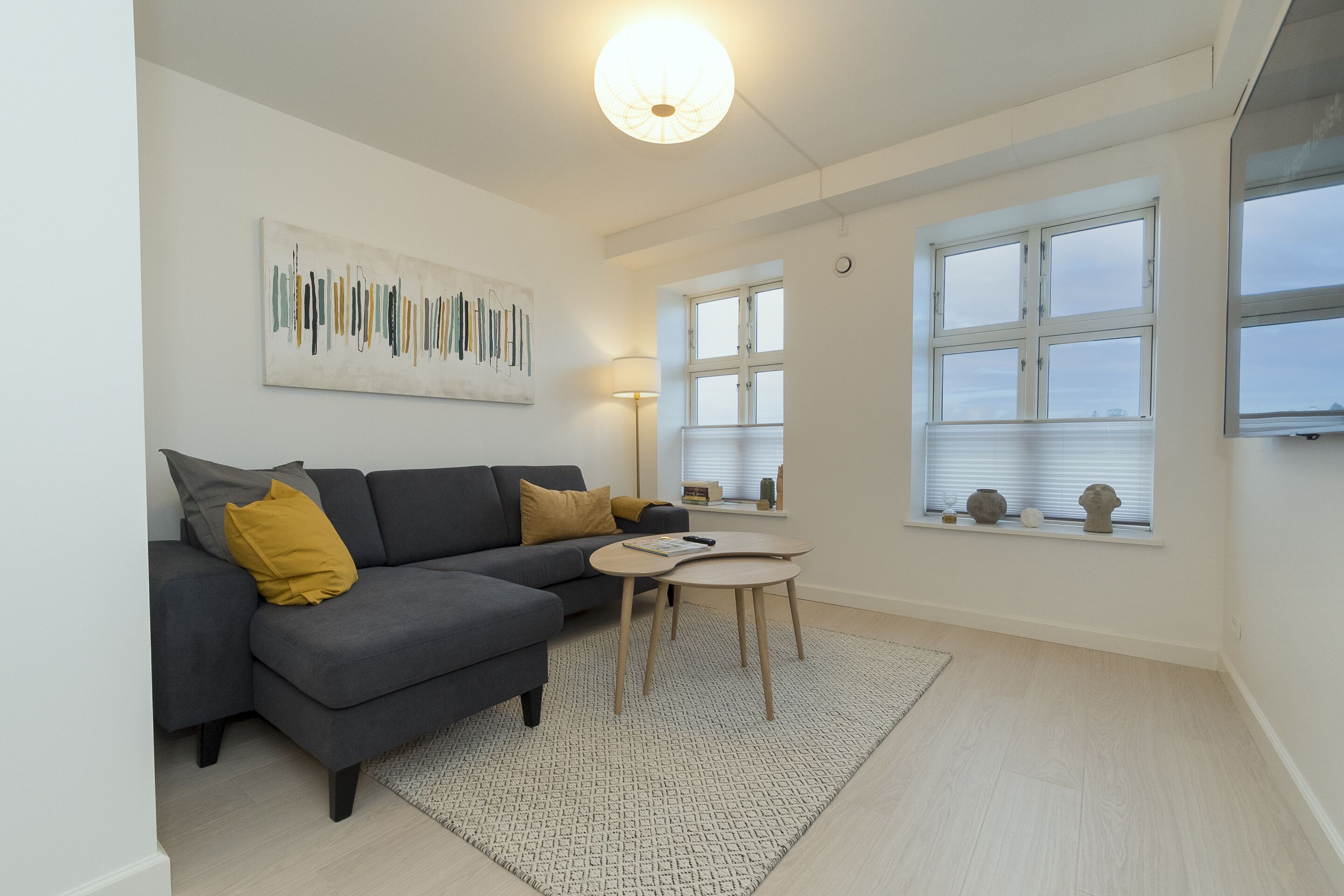 Ein modernes Wohnzimmer mit einem grauen Ecksofa, einem kleinen runden Tisch und einem großen Fenster mit Jalousien.