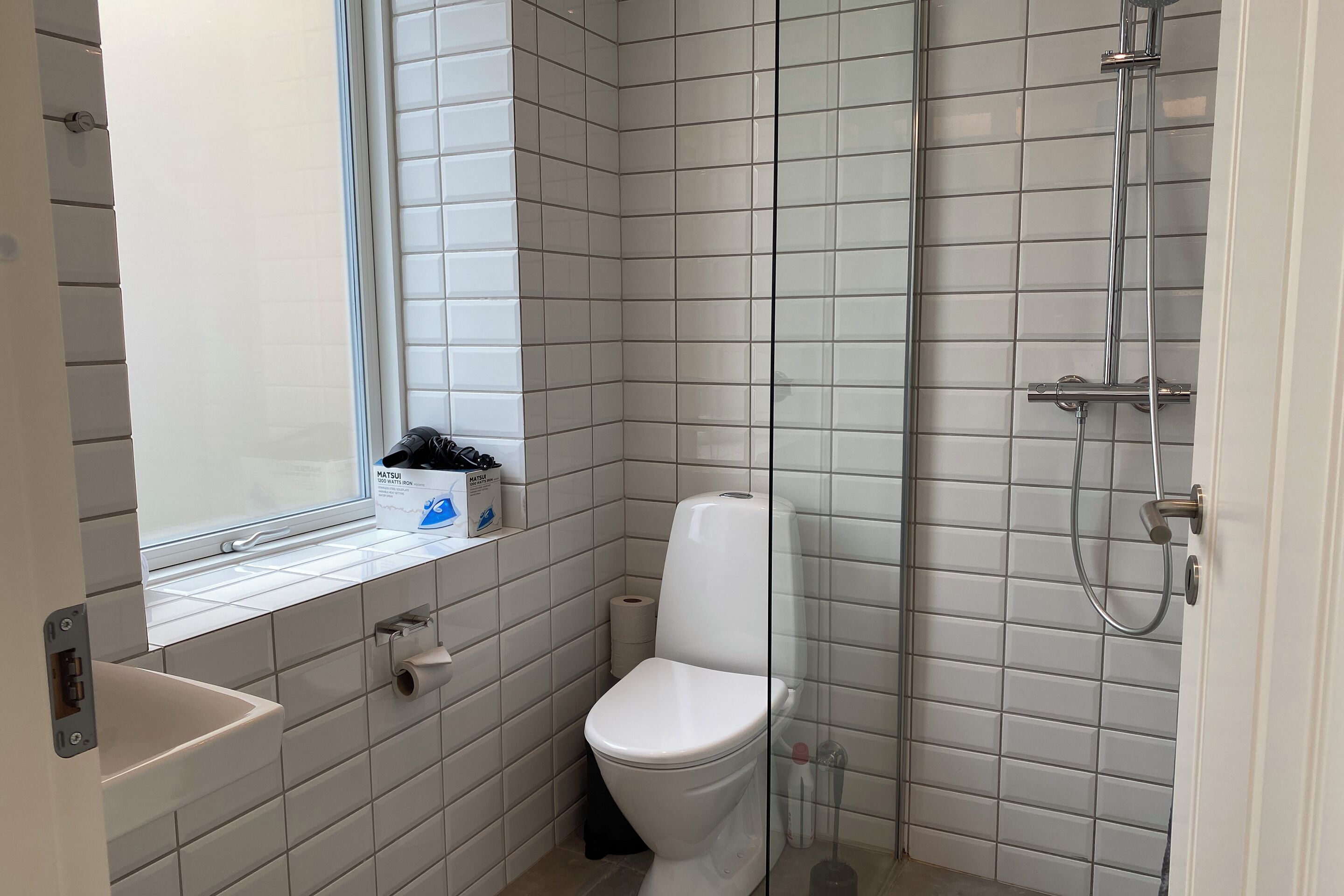 Ein kleines, sauberes Badezimmer mit weißen Fliesen, einer Toilette, einer Dusche und einem Fenster.