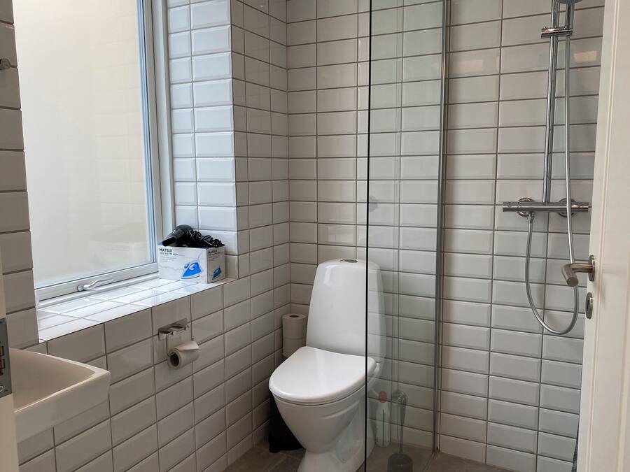 Un baño pequeño y limpio con azulejos blancos, un inodoro, una ducha y una ventana.