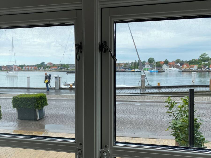 Vista pela janela de um porto com barcos, um pedestre e edifícios ao fundo.