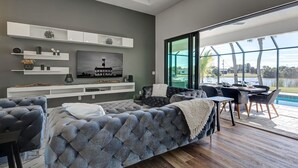 Ein modernes Wohnzimmer mit grauem, gestepptem Sofa, einem Flachbildfernseher auf einem weißen Regal und einem Essbereich mit Sitzgelegenheiten im Freien neben einer Glasschiebetür mit Blick auf einen See.