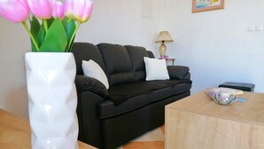 Un salon avec un canapé noir, un vase blanc contenant des fleurs roses, une lampe et une petite table sur laquelle se trouvent un livre et un objet décoratif.