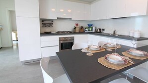 A modern kitchen with white cabinets, a gas stove, an oven, and a microwave. There is a dining area with a table set for two, featuring plates, glasses, and cutlery. - The Green House on the Lake (Lezzeno)