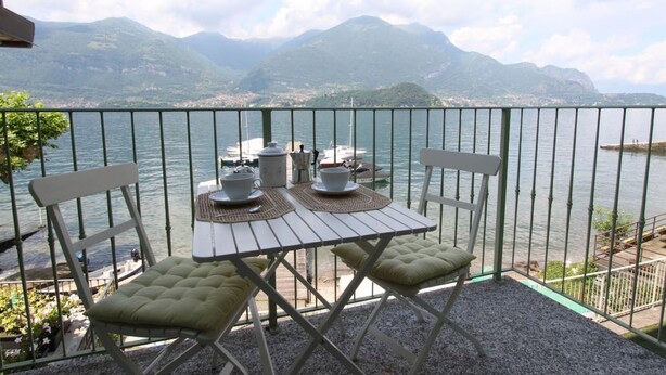 A balcony with a table set for two, overlooking a lake with boats and mountains in the background. - The Green House on the Lake (Lezzeno)