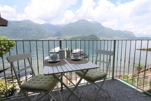 A balcony with a table set for two, overlooking a lake with boats and mountains in the background.