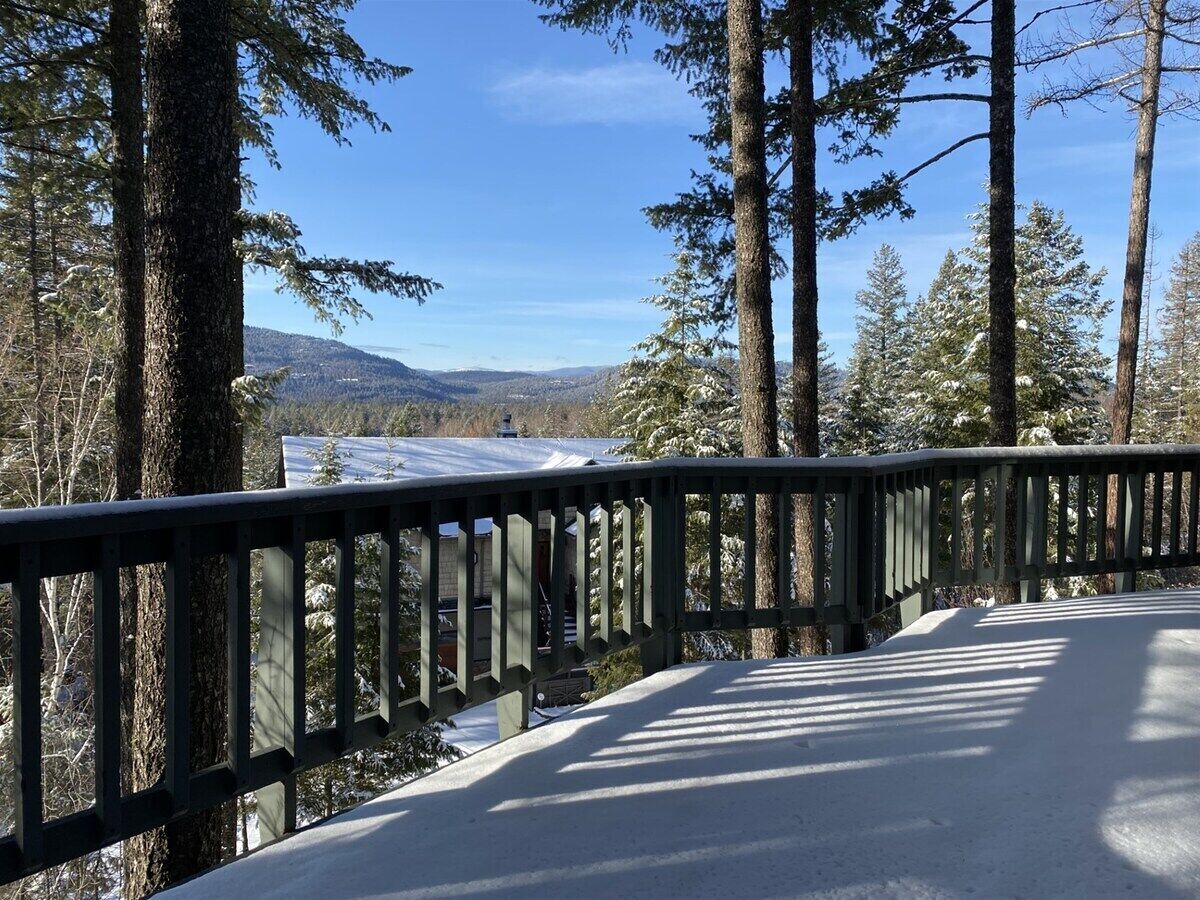 En snötäckt terrass med ett räcke, omgiven av höga, vintergröna träd och med en fri utsikt över ett avlägset landskap.