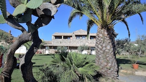 Un bâtiment en pierre, surmonté d’un toit en tuiles, entouré d’un jardin bien entretenu où l’on trouve des palmiers et des cactus.