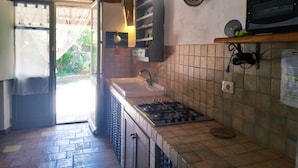 Une cuisine équipée d’une cuisinière à gaz, d’un évier et d’un plan de travail recouvert de carreaux. On y trouve une fenêtre avec des rideaux, un luminaire fixé au mur et une étagère contenant des ustensiles de cuisine.
