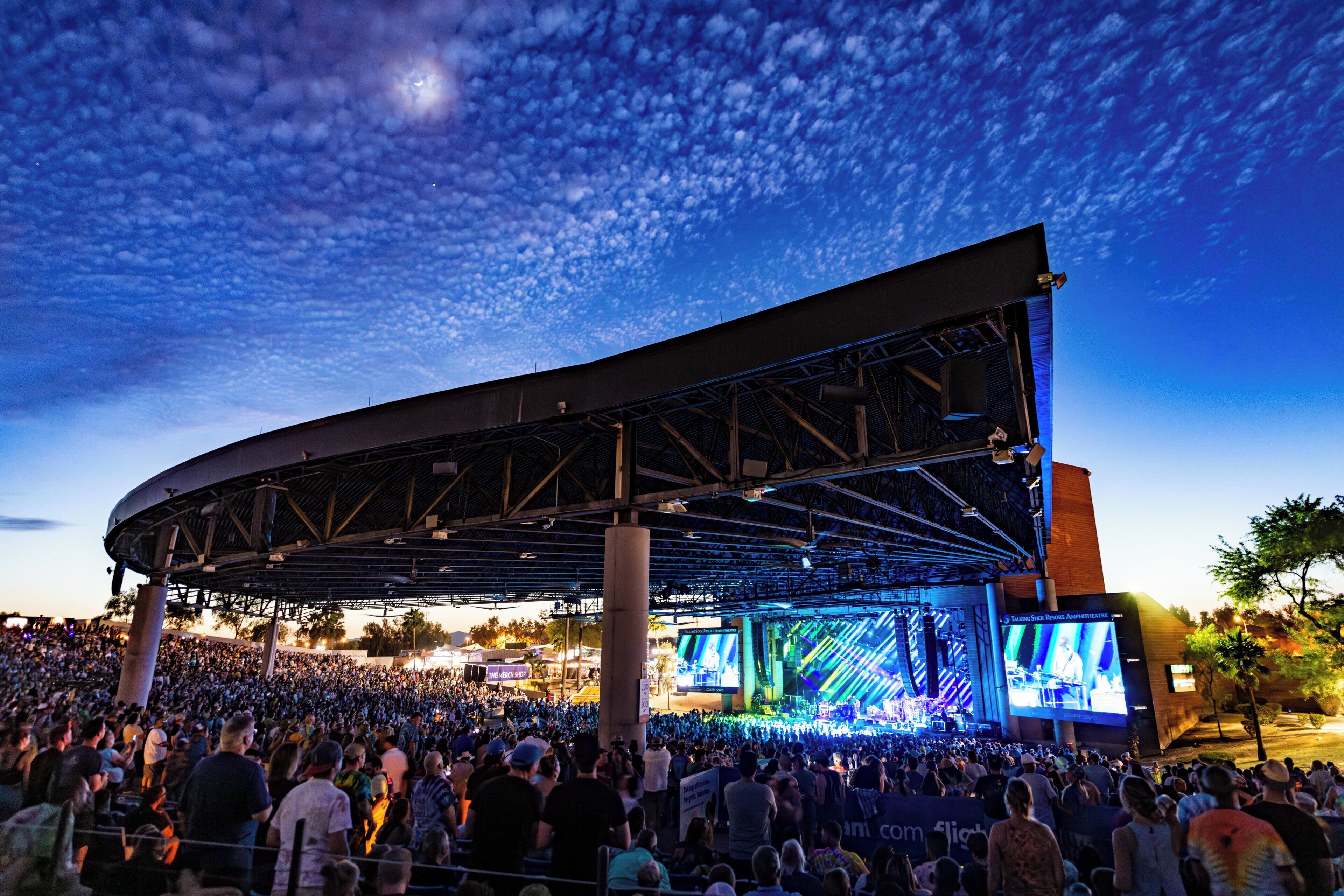 A large outdoor amphitheater with a curved stage and multiple screens, surrounded by a crowd of spectators.