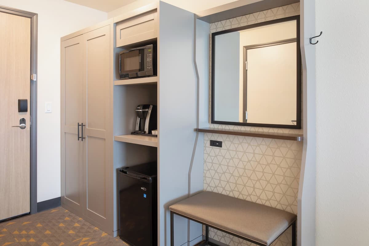 A compact hotel room with a wardrobe, a microwave, a coffee machine, and a small seating area.