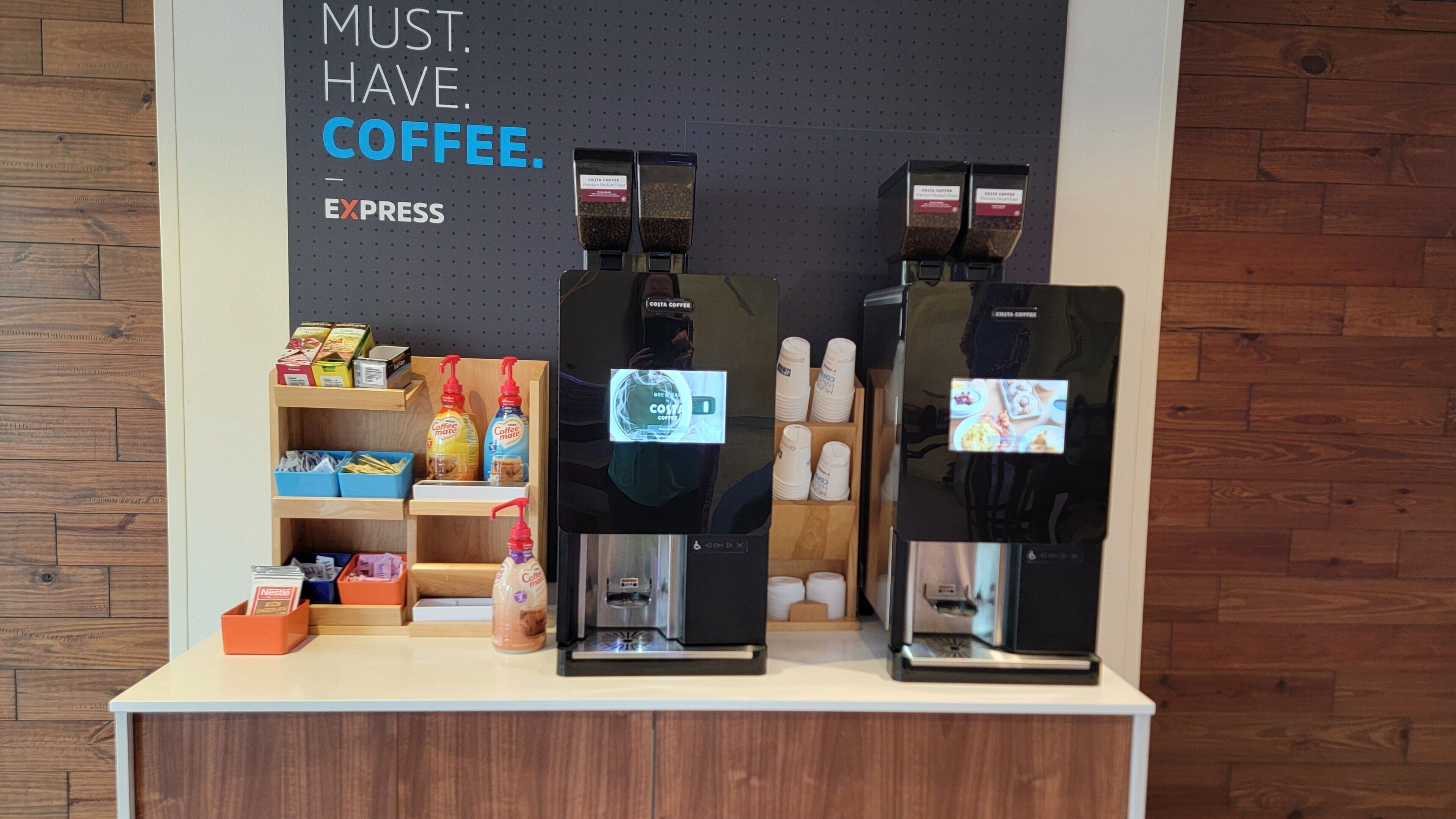 A coffee station with two espresso machines, a shelf with coffee products, and a sign that reads 'MUST. HAVE. COFFEE.'