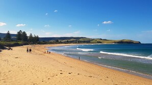 Ein weiter Sandstrand mit sanften Wellen, einem klaren Himmel und Hügeln in der Ferne.