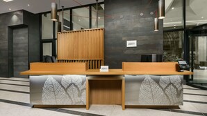 En receptionsdisk med modern design, med en trätopp och en metallbas som har ett bladmotiv. Två hänglampor är placerade ovanför disken, och en liten skylt visar hotellets namn.