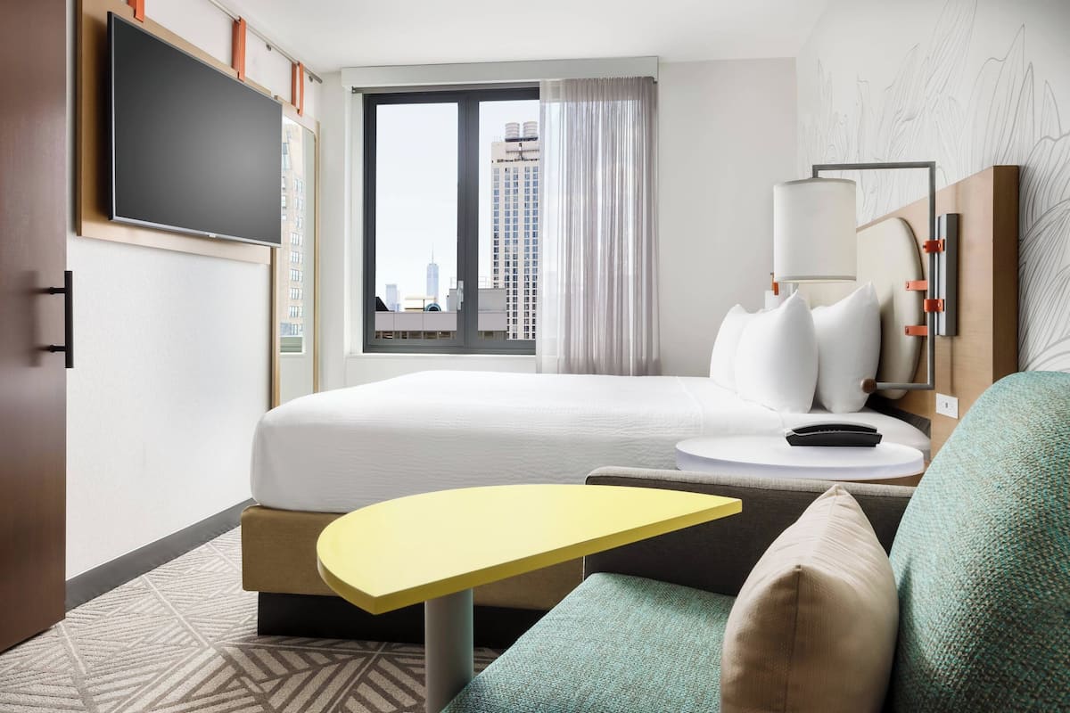 Una habitación de hotel con una cama, una mesita amarilla pequeña, un sofá, un televisor montado en la pared y una ventana con vistas a la ciudad.