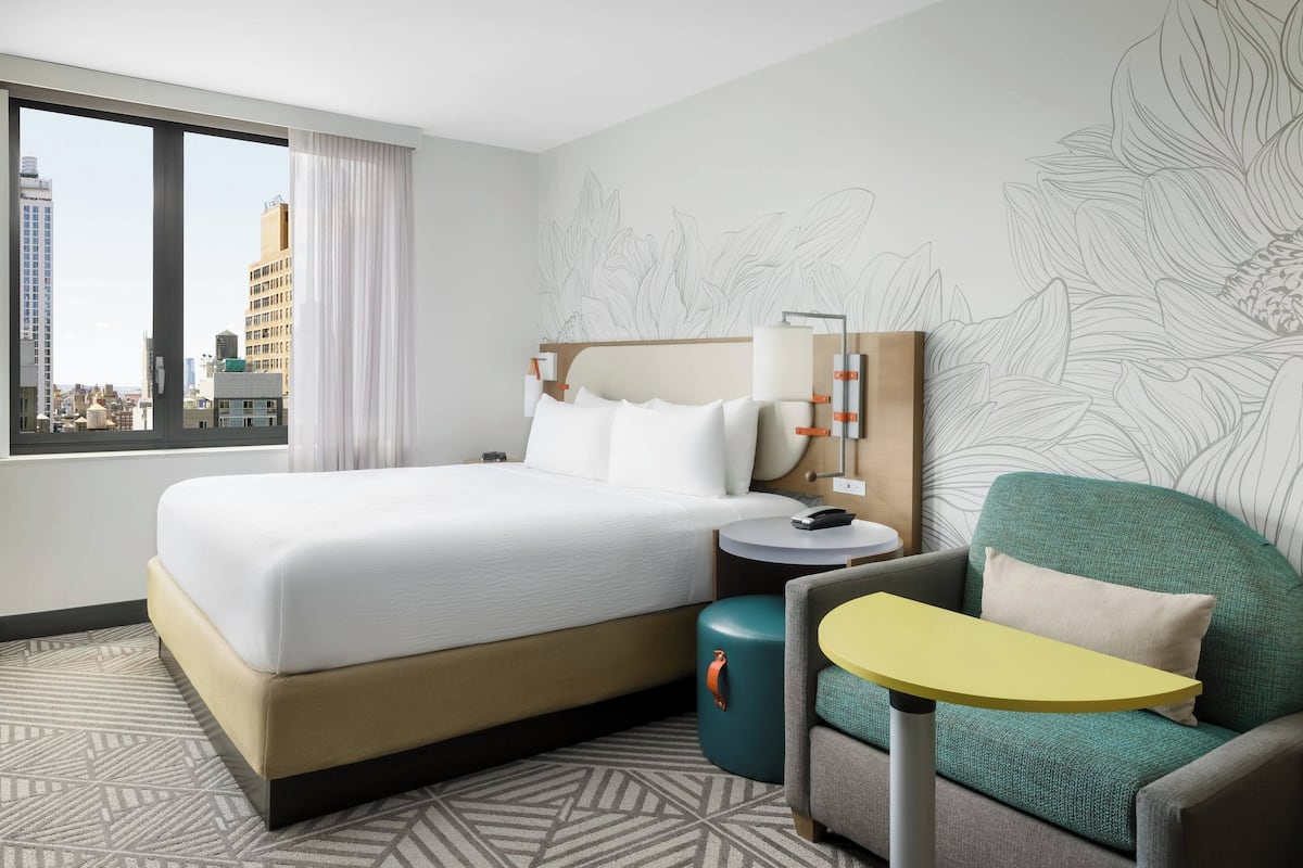 Habitación de hotel con una cama grande, una mesita redonda pequeña, un sillón verde y vistas de la ciudad a través de un ventanal amplio.