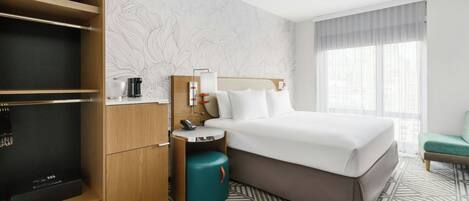 A hotel room with a bed, a desk with a lamp, a minibar, and a large window.