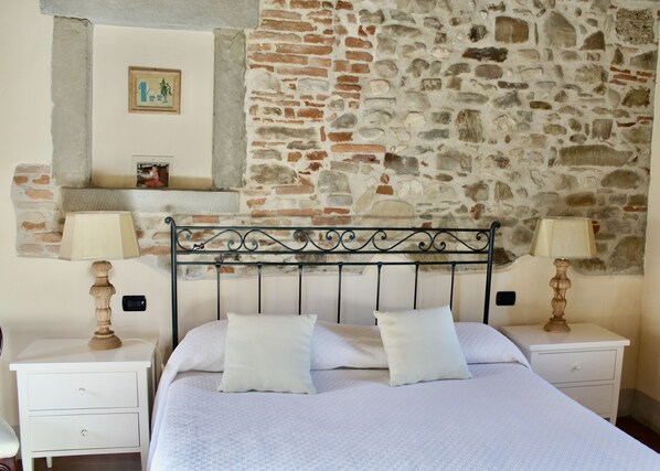 A bedroom with a stone wall, a bed with white bedding, two bedside tables with lamps, and a small shelf with a picture.