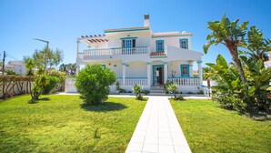 Casa de dos pisos con fachada blanca y detalles azules, entrada principal y camino de acceso pavimentado.