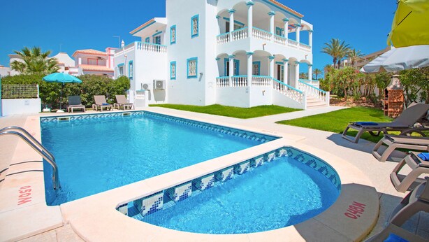 Una piscina con una característica de agua azul cristalina, rodeada de sillones de descanso y sombrillas, y un edificio blanco con balcones al fondo.