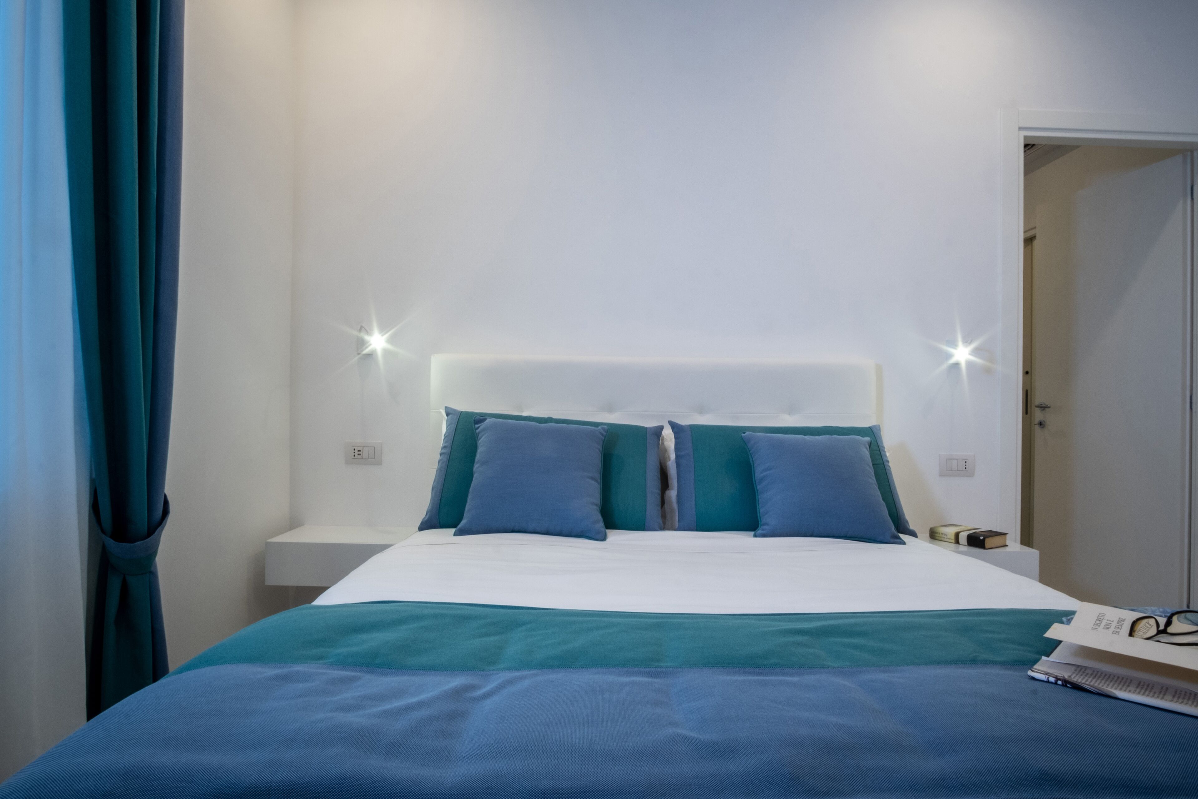 Ein ordentlich bezogenes Bett mit weißem Kopfteil und blaustichigem Bettwäsche, ein Nachttisch mit Lampe sowie eine Tür zu einem anderen Raum.