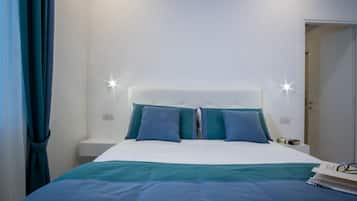 Een netjes opgemaakt bed met een wit hoofdbord en blauwgroene beddengoed, een nachtkastje met een lamp, en een deur die naar een andere kamer leidt.